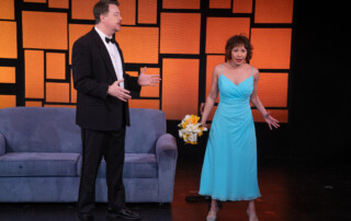 A man in a suit stands beside an exasperated bridesmaid in a teal dress with a small off-white bouquet of flowers.