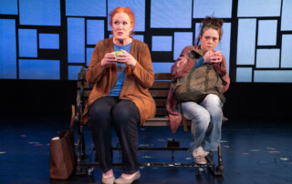 An older woman with red hair sits on a park bench with a young woman cradling a backpack.