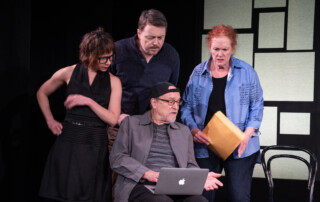 A woman, man, and older woman stand around an older man with a laptop, peering at his screen.