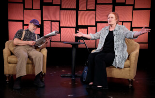 An older man looks at his newspaper while sitting in an armchair next to a woman sitting in an identical chair waves her hands around animatedly.