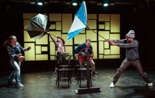 A young woman looks nervous with her hair flying in the wind. Next to her, a woman with an inside-out umbrella held above her head seems to struggle to keep her feet on the ground. Next to her, a man crouches from an unseen force. In the foreground, a young man holds an inside-out umbrella at an angle, bracing himself against an unseen wind.