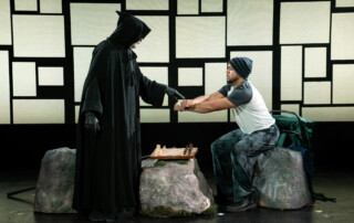 A man dressed as the grim reaper holds his hand out to a young man sitting on a rock. A chess board sits on another rock between them.