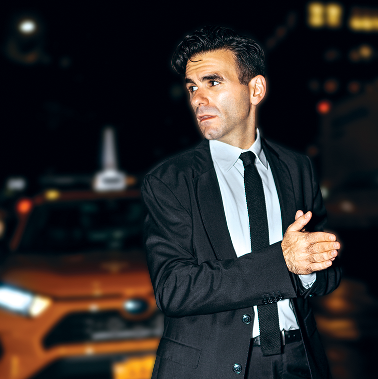 A dark-haired man looks off camera with his hands pressed together. He wears a black suit and stands in front of a yellow taxi.