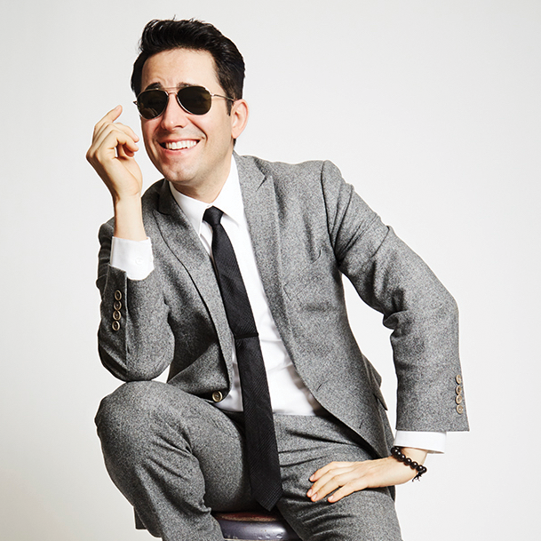 JohnLloydYoung_1x1 A man in a gray suit and black tie casually sits on a stool with a smile on his face and a pair of sunglasses atop his nose.