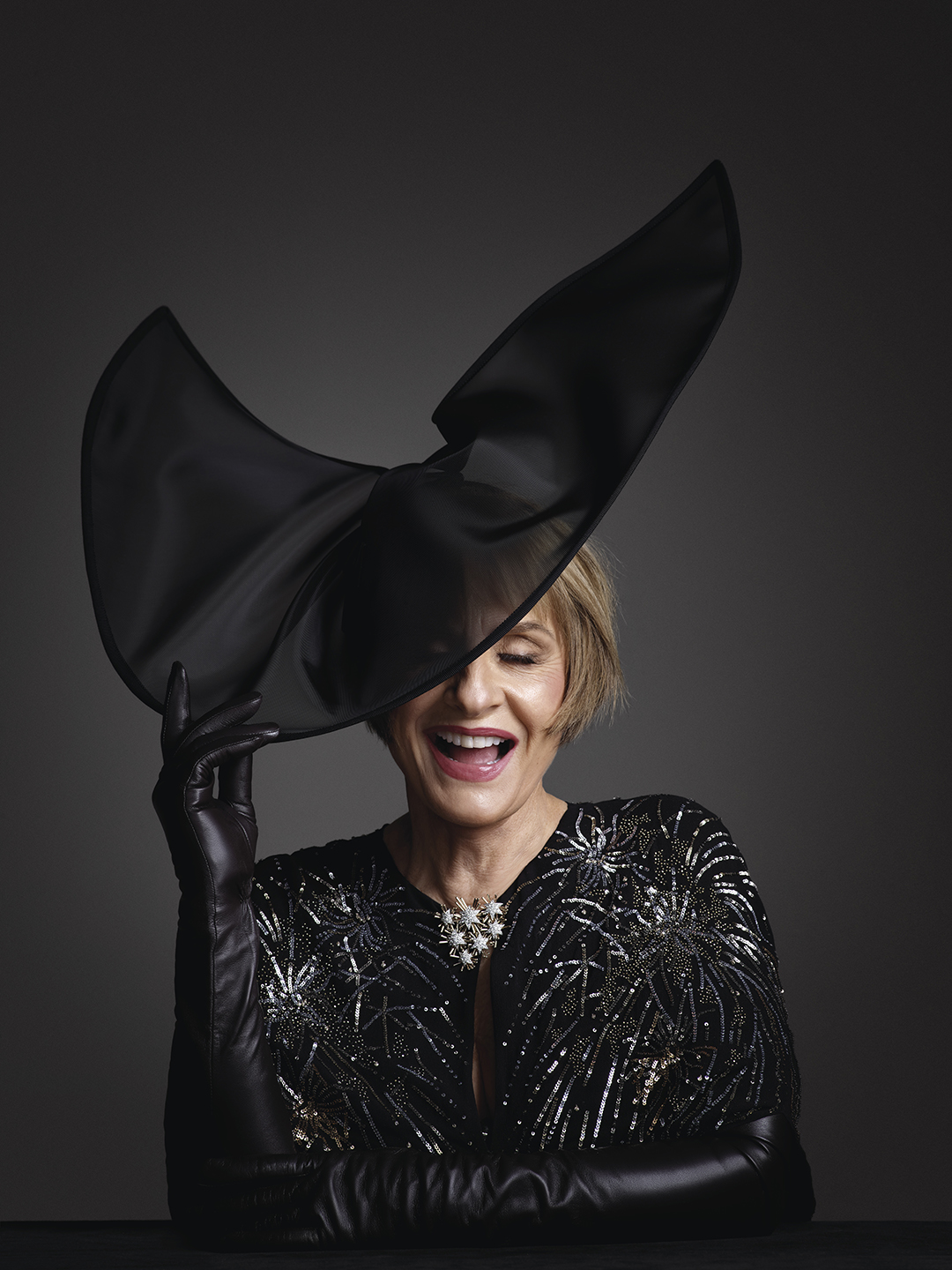 A laughing woman with cropped, light brown hair poses with a wide, semi-opaque black hat between her fingers. The brim of the hat covers her right eye. She wears elbow-length black leather gloves and a sequined black gown with a brooch made of a cluster of silver stars.